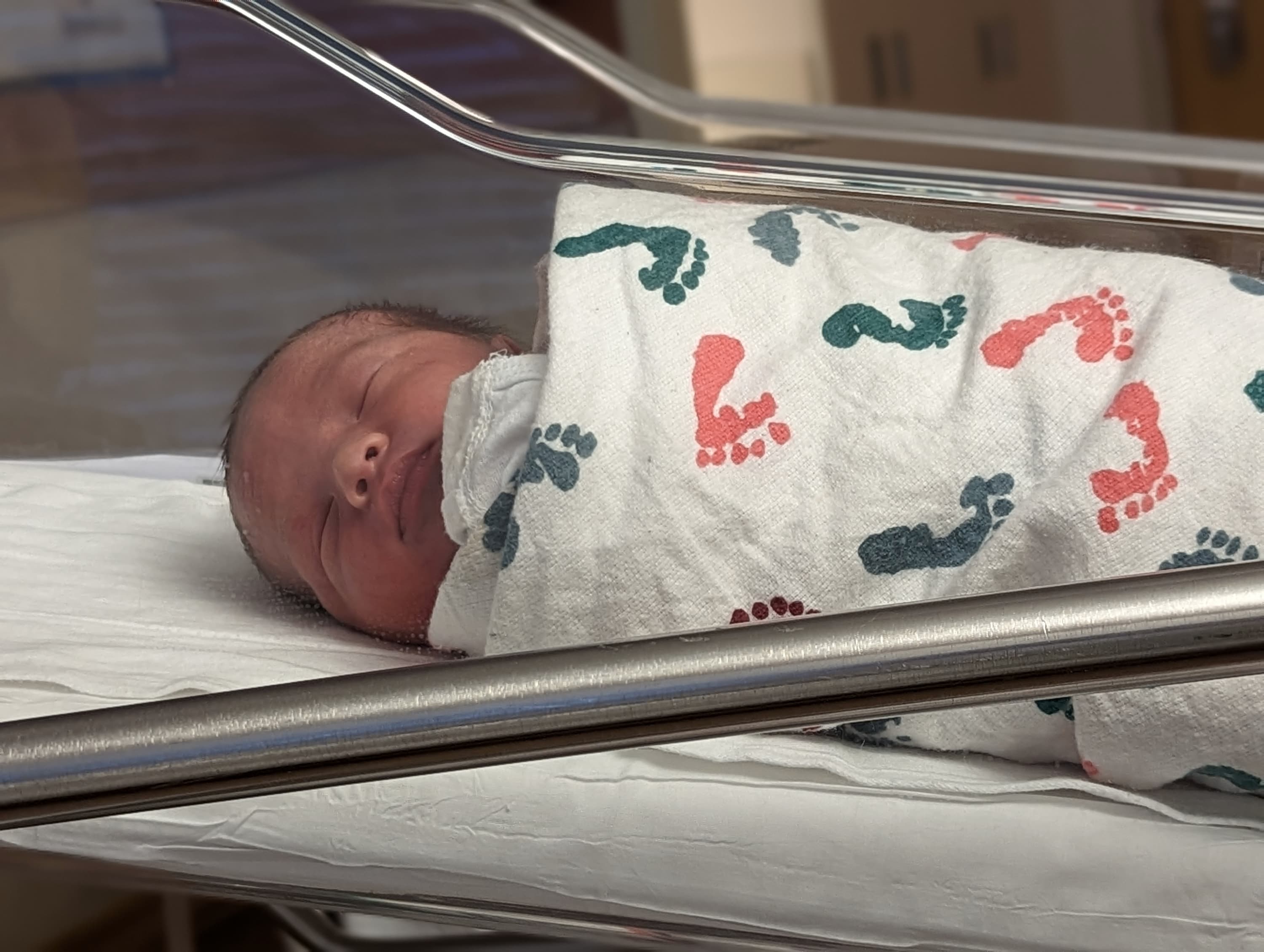 A baby asleep in a bassinet, swaddled in a white blanket with different colored footprints on it.
