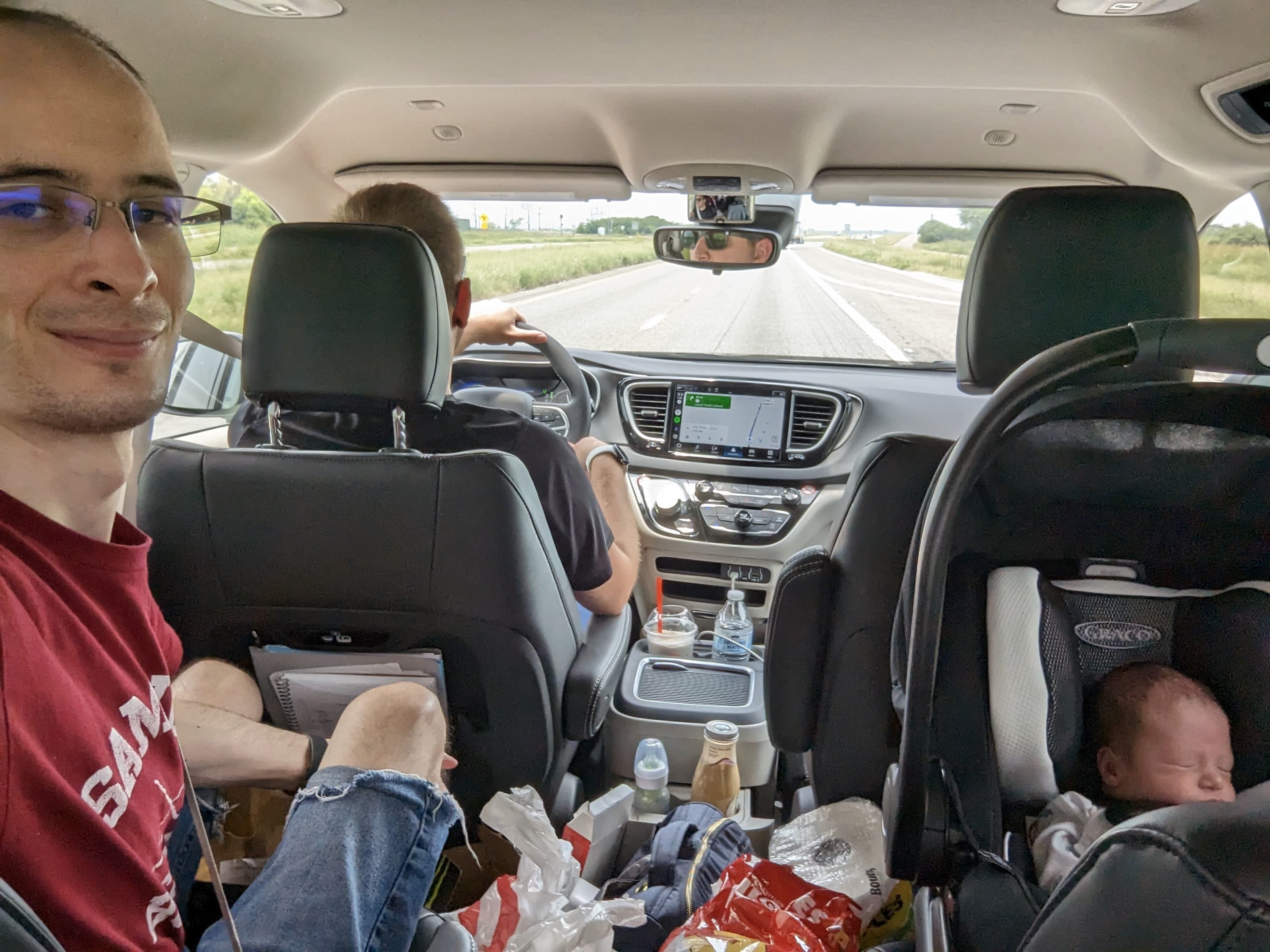 Paddy taking a selfie in the bucket seat in the back of a minivan. A baby is sleeping in a carseat in the other bucket seat. Ethan is driving in the front seat. The back of the minivan is littered with bags and drinks.