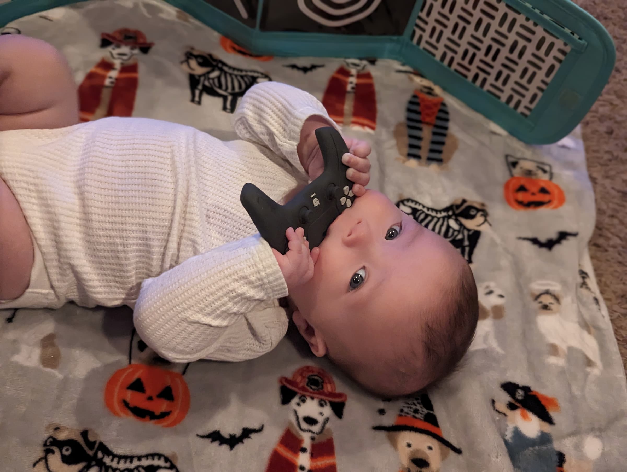 A baby looking at the camera, laying on its back. It has a teether toy shaped like a controller that it's holding in its mouth.